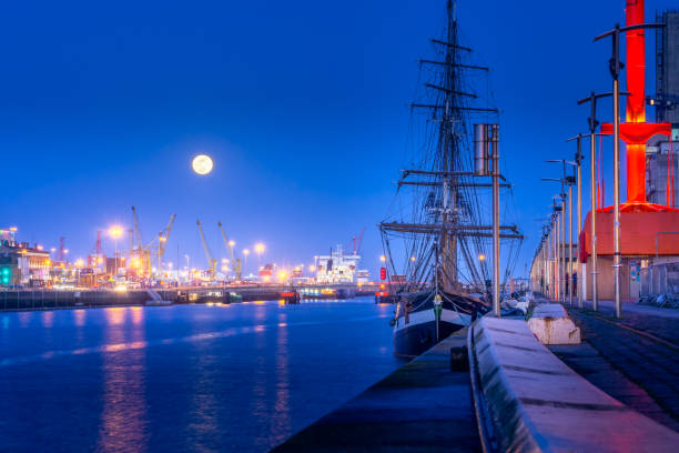 Tall ship docked in Dublin dock on Liffey river, blue hour and full moon. Illuminated embankment and blurred water, long exposure photography, Ireland