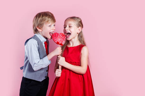 心の形のキャンディレッドロリポップを持つ面白い男の子と小さな女の子。美しい子供たちはお菓子を食べる。バレンタインデー - child valentines day candy eating ストックフォトと画像