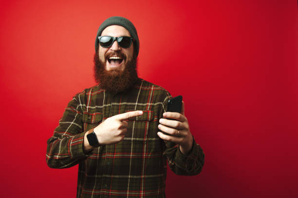 istock Cheerful amazed bearded hipster man pointing at phone over red background 1199748902