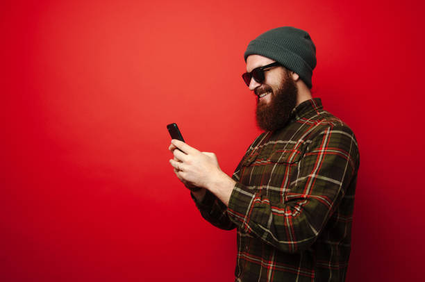 istock Excited young bearded hipster man using smartphone over red background 1199749413