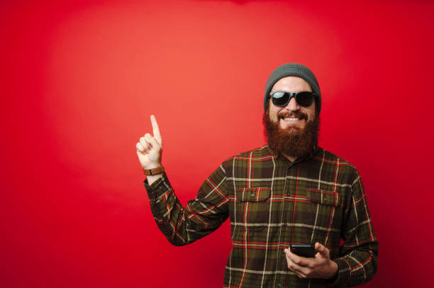 istock Young happy bearded man holding phone and pointing up at copyspace 1199749431
