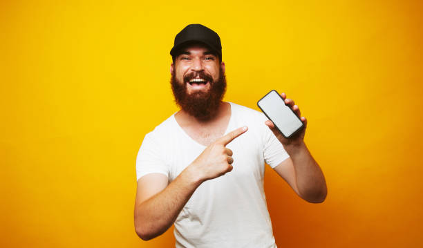 istock Happy bearded hipster man pointing at smartphone over yellow background 1199749811
