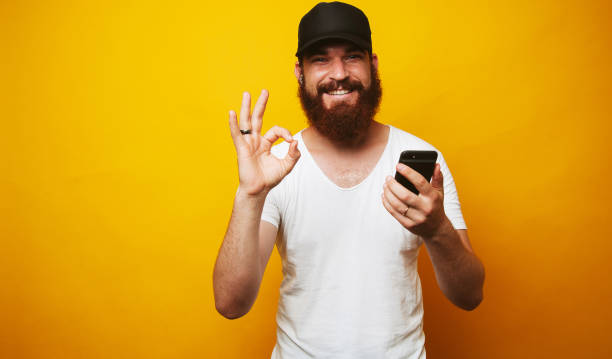 istock Man with beard holding phone and showing OK gesture 1199749902