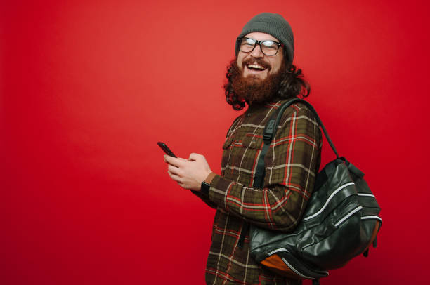 istock Cheerful smiling hipster man with beard using phone and wearing backpack over red background 1199751862