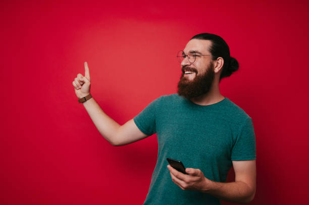 istock Happy hipster man with beard holding smartphone and pointing away at copyspace over red background 1199751978