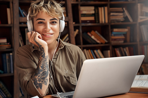 Portrait of happy and charming young adult woman spending day at university library with books on bookshelves. Smiling girl with headset sitting behind table with laptop and looking at camera