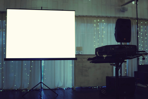 Empty projection screen put in the centre of a illuminated stage in a banquet hall. Projection screen equipment at the decorated curtain background. Facilities for video projection at a festive event