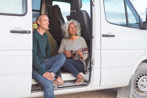 An adorable mixed-race senior couple sit in the open doorway of their camper van and make music together. She is playing the ukulele.
