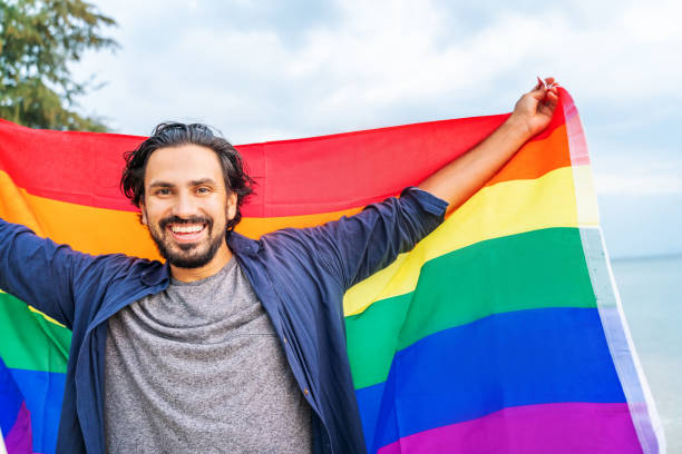 un tipo alegre con una bandera arco iris en la playa. joven sosteniendo una bandera arco iris contra el cielo del océano - gay pride rainbow flag homosexual fotografías e imágenes de stock