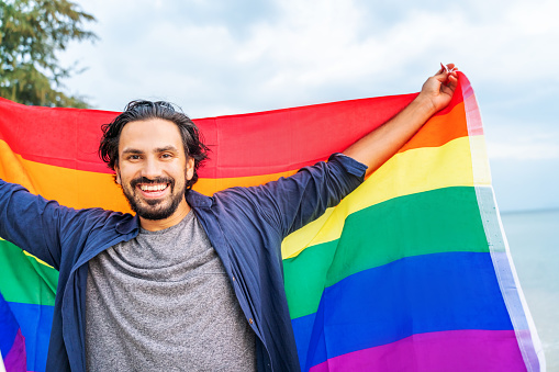 Cheerful latin guy with a rainbow flag on the beach. Young man holding a rainbow flag against the ocean sky