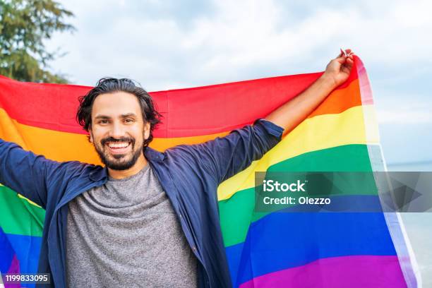 Cheerful Guy With A Rainbow Flag On The Beach Young Man Holding A Rainbow Flag Against The Ocean Sky Stock Photo - Download Image Now