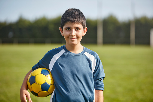 Close-up of 12 year old Spanish male footballer in blue jersey holding ball under arm and smiling at camera.