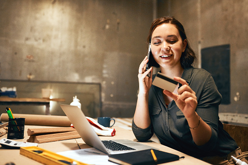 Cropped shot of an attractive young female carpenter holding her credit card and doing online shopping while talking on her cellphone inside of a workshop