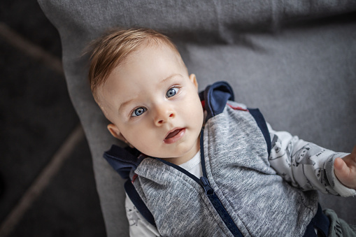 Close up of adorable toddler with big blue eyes lying on gray background and looking at camera.