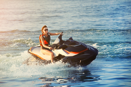 Young adult sporty caucasian woman riding jet ski in ocean blue water at warm evening sunset. Beach extreme sport activities and recreation.