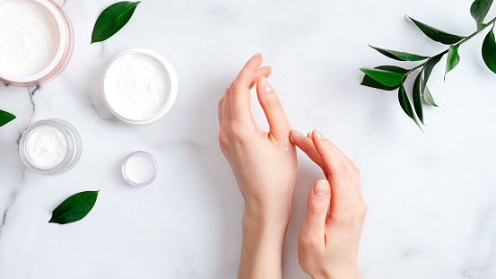 Cosmetic cream on female hands, jars with milk swirl cream and green leaves on white marble table. Flat lay, top view. Woman applying organic moisturizing hand cream. Hand skin care concept