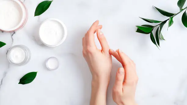 Photo of Cosmetic cream on female hands, jars with milk swirl cream and green leaves on white marble table. Flat lay, top view. Woman applying organic moisturizing hand cream. Hand skin care concept