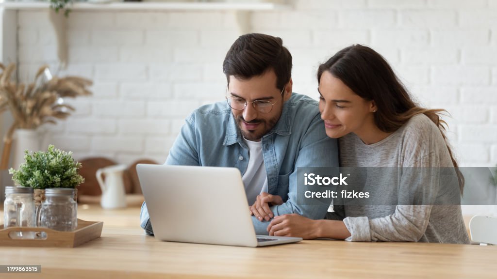 Happy young mixed race married spouse making purchases online. Pleasant family couple sitting at big wooden table in modern kitchen, looking at laptop screen. Happy young mixed race married spouse web surfing, making purchases online or booking flight tickets. Couple - Relationship Stock Photo