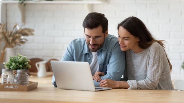 Happy young mixed race married spouse making purchases online. Pleasant family couple sitting at big wooden table in modern kitchen, looking at laptop screen. Happy young mixed race married spouse web surfing, making purchases online or booking flight tickets. wife stock pictures, royalty-free photos & images