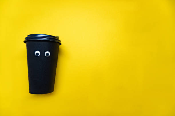 istock Black coffee paper cup with cap with googly plastic eyes are on yellow background, enough space for the text. 1199864525