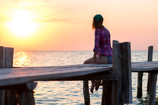 the girl is sitting on the old wooden berth and admires sunset at sea