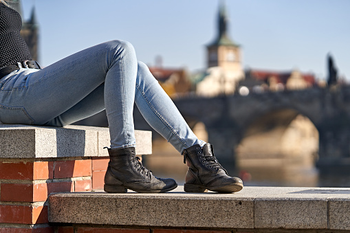 Close-up of a female traveler sit down on a stone wall and dressing casual jeans and to and old style leather black boots with a blurred background of the Bridge of Charle in Prague. Holidays concept.