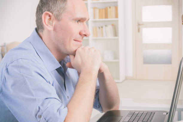 A man is deeply engaged in work using his laptop, paired with a notebook for planning. The scene captures focus and determination in a dimly lit home office setting.