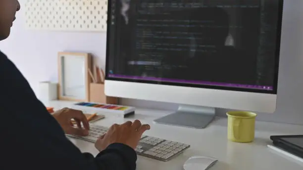 Photo of Cropped shot from back view of young programmer while he is typing/coding on the keyboard in front of the monitor at the working desk.