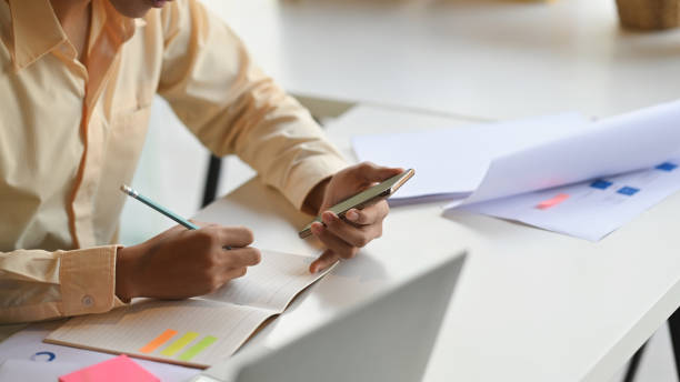 istock Cropped shot of young businessman while using cellphone and sitting at the work desk. Writing and looking at phone. Focus on hand with phone. 1199885367