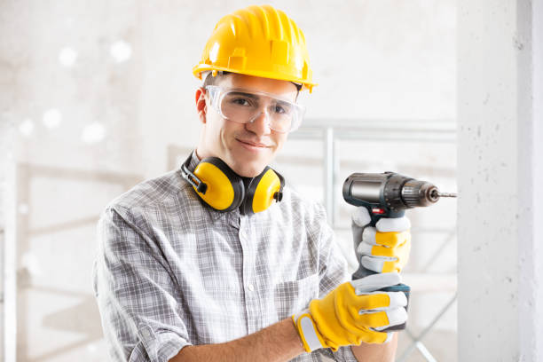 Young construction worker drilling wall indoors Young construction worker wearing yellow hardhat, gloves and safety goggles drilling a wall inside new build house, holding electric drill tool with both hands, looking at camera and smiling drill photos stock pictures, royalty-free photos & images