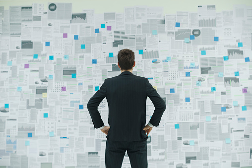 Corporate businessman standing with arms akimbo in front of a wall with lots of financial reports, business strategies and planning concept