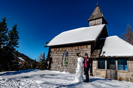 An elderly woman stands next to a snowman in front of Mountain church in the European Alps, Austria, Nassfeld, Pramollo,Nikon D850