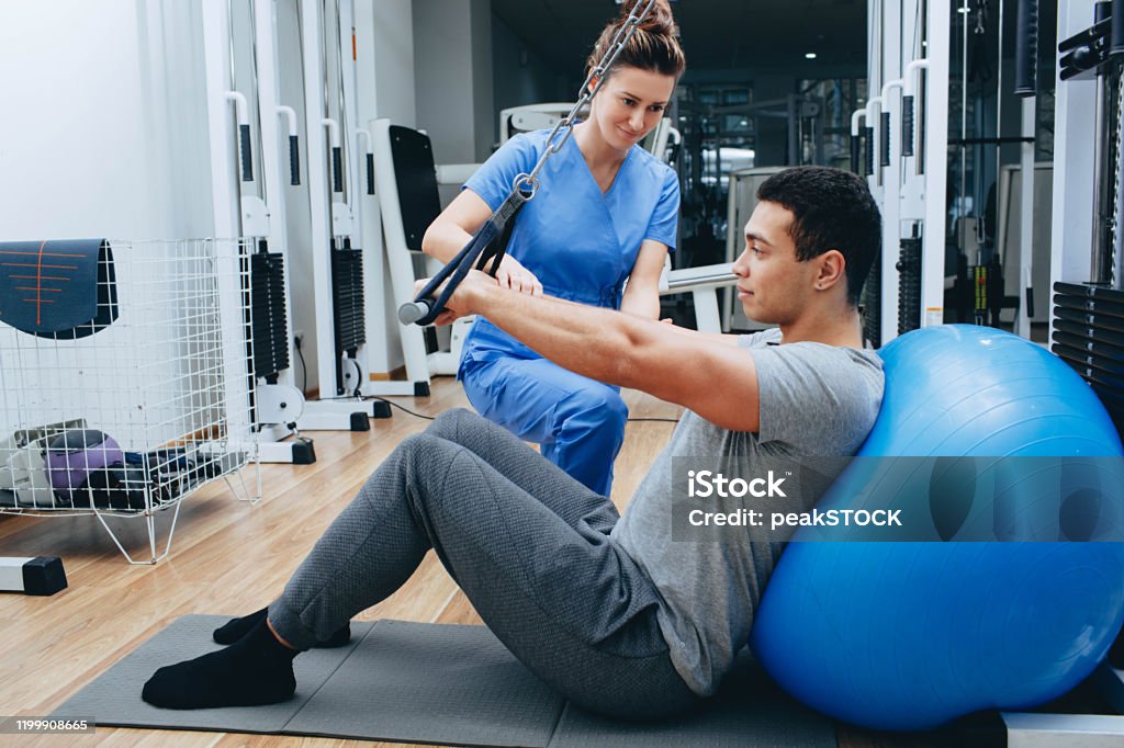 kinesiologist helps a mixed race man doing exercises to strengthen his back muscles. treatment of back pain using kinesitherapy. Physical Therapy Stock Photo