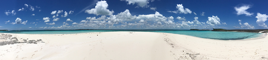 Panoramic image of white sand and turquoise waters underneath a bright blue sky with white puffy clouds in the Sea of Abaco in the Bahamas; landscape