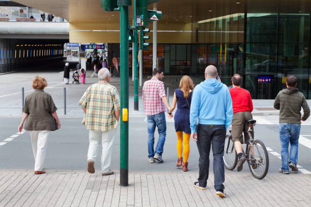 People are walking over crosswalk at main station Essen and they are coming from square Freiheit opposite to station. There people different ages and ethnicities. A man is cycling. A couple is walking hand in hand. Man is watching at woman while walking.