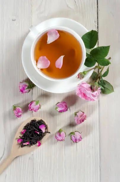 Cup of tea, rosebuds and wooden spoon with tea on  white wooden table with copy space. Top view.