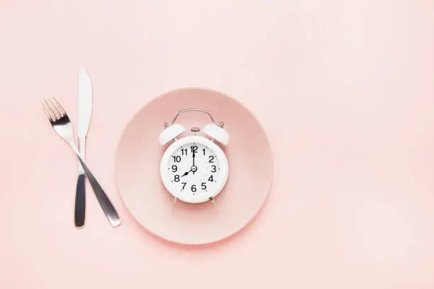 Intermittent fasting concept. White alarm clock on empty pink dish with knife and fork on pink background. Flat lay, copy space for text.