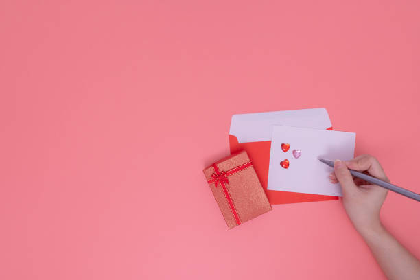istock love concept, a right-hand holds a pen write on a white paper. that put on a red envelope and red gift box beside. there are shiny hearts put on a corner of a paper. Those are put on a pink background 1199925378