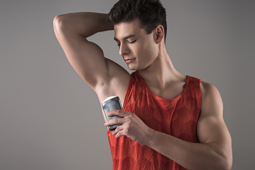 young man in red sleeveless shirt applying deodorant on underarm isolated on grey