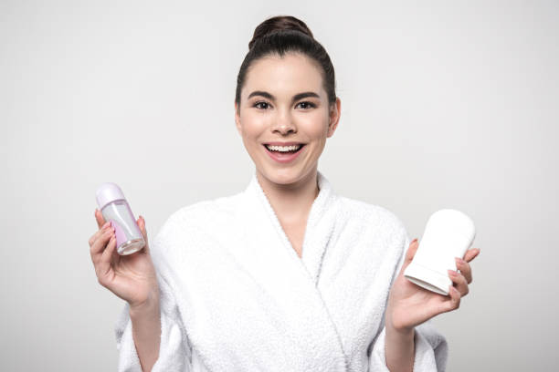 istock cheerful woman in bathrobe holding deodorants while looking at camera isolated on grey 1199927943
