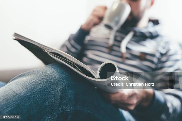 Man Reading Magazine Or Newspaper And Drinking Coffee While Relaxing On Sofa Stock Photo - Download Image Now