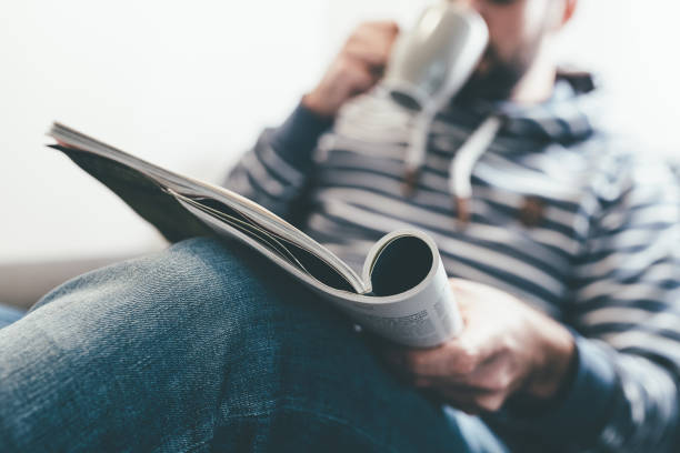 man reading magazine or newspaper and drinking coffee while relaxing on sofa - news magazine imagens e fotografias de stock