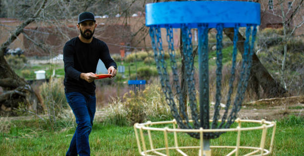 a man in his thirties with a beard prepares to throws a disc golf putter into a hanging disc golf basket next in a park outdoors - golf expertise professional sport men imagens e fotografias de stock