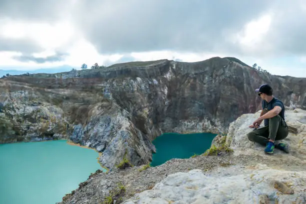 Man sitting at the volcano rim and watching the Kelimutu volcanic crater lakes in Moni, Flores, Indonesia. Man is relaxed and calm, enjoying the view on lake shining with many shades of turquoise