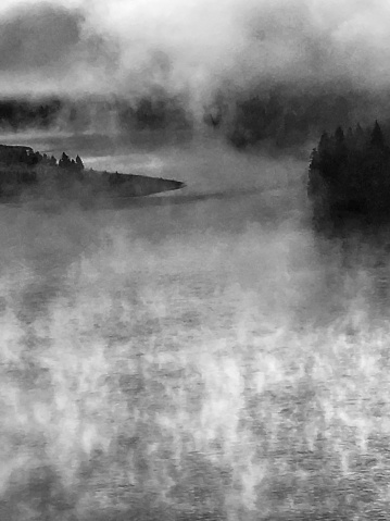 A black and white scenic abstract landscape of fog mist rising from the Peace River in British Columbia, Canada, with forest trees on islands in the river water.