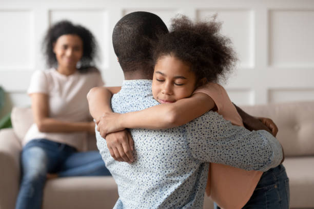 Little school girl embracing mixed race father. Young african american man holding, embracing, comforting smiling happy calm black cute kid daughter, blurred mother sitting on couch on background, loving supporting family concept. mercy hug stock pictures, royalty-free photos & images