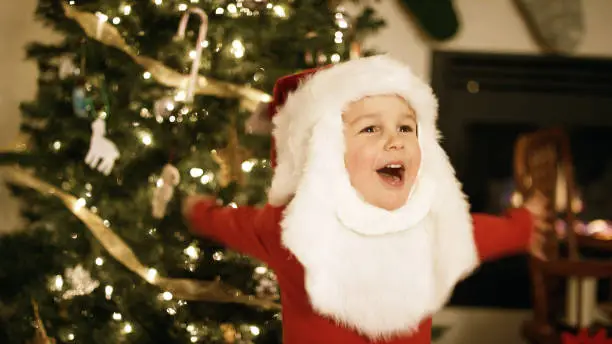 A Three Year-Old Caucasian Boy Laughs while Wearing a Santa Claus Hat and Beard in Front of a Christmas Tree