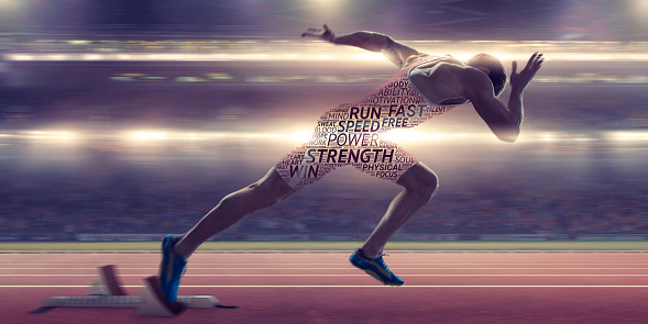 A conceptual image of a male athlete sprinting out from the starting blocks on a running track in a floodlit stadium. His body is covered with a motivational word cloud with positive words in differing fonts and sizes. With motion blur.