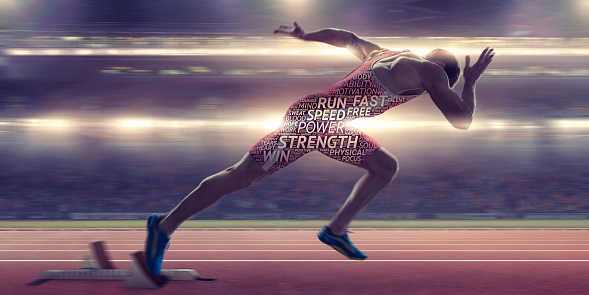 A conceptual image of a male athlete sprinting out from the starting blocks on a running track in a floodlit stadium. His body is covered with a motivational word cloud with positive words in differing fonts and sizes. With motion blur.