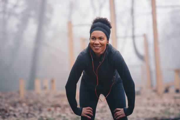 młoda afrykańska kobieta odpoczywa po sesji joggingu w parku. uśmiecha się. stoi przed sprzętem do ćwiczeń na świeżym powietrzu. nosi słuchawki. to zimny, mglisty, jesienny dzień. - hand on knee audio zdjęcia i obrazy z banku zdjęć